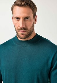 Turkusowy sweter z okrągłym dekoltem, wykonany z dzianiny, z prążkowanym wykończeniem dekoltu i miękką fakturą. Zbliżenie pokazujące szczegóły i głębię koloru.