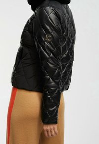 Svart quiltad puffjacka med en glänsande yta, med en rund logotyp på vänster ärm, hög krage och en kortad design.