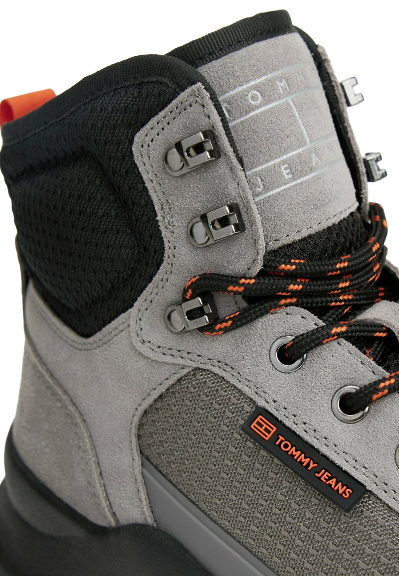 Graue Wildleder- und Netzstiefel mit schwarzen Akzenten, ausgestattet mit Metall-D-Ringen, orangefarbenen Schnürsenkeln und einem rechteckigen Logo-Patch. Dicke Sohle für Dämpfung.