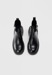 Mustat nahkaiset nilkkasaappaat, joissa on sileä pinta, jykevät mustat kumipohjat, sivuvetoketjut ja takana lenkkivetimet.