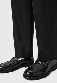 Black leather loafers with textured toe and glossy finish, paired with black trousers. Features a decorative strap across the instep.