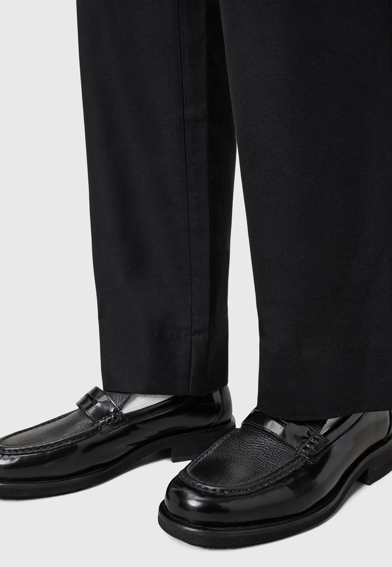 Black leather loafers with textured toe and glossy finish, paired with black trousers. Features a decorative strap across the instep.