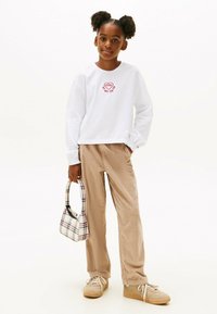 Weißes Langarmshirt mit roter Herzstickerei, beigem Cordhosen und einer karierten Handtasche. Beige Sneakers mit weißen Schnürsenkeln.