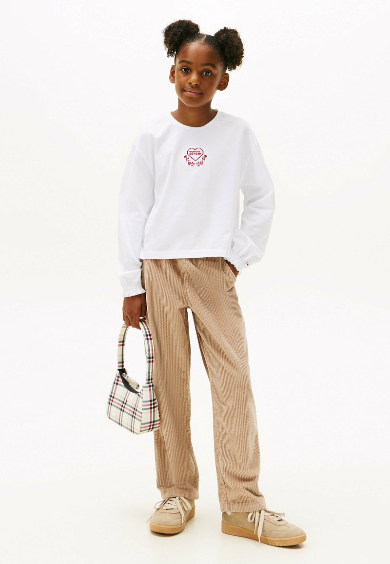 Weißes Langarmshirt mit roter Herzstickerei, beigem Cordhosen und einer karierten Handtasche. Beige Sneakers mit weißen Schnürsenkeln.