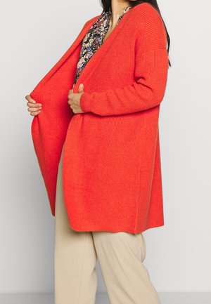 Frau trägt eine knallorange gestrickte Strickjacke über einer geblümten Bluse und beigen Hosen, hält die Strickjacke mit beiden Händen geöffnet.