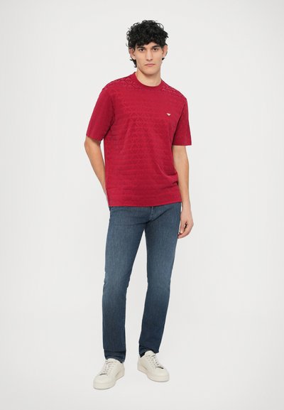 Rdeča kratka majica s teksturiranim vzorcem in okroglim izrezom, kombinirana z modrimi kavbojkami in belimi supergami. Preprost, vsakdanji dizajn.