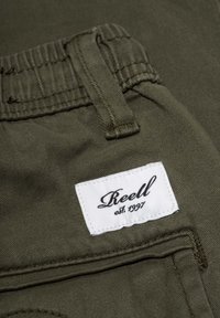 Pantalones verde oliva con una tela suave y texturizada. Presentan una cinturilla elástica y una etiqueta blanca que dice "Reell est. 1997" cosida en el lateral.
