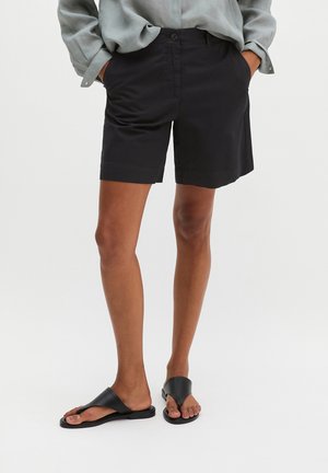 Persoon die zwarte knielange shorts draagt, lichtgrijs shirt met lange mouwen met handen in de zakken, en zwarte instapsandalen op een witte achtergrond.