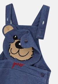 Blaue Denim-Overalls mit einer Bärenkopf-Stickerei in Beige und Schwarz, rotem Text "guess" und verstellbaren Trägern.