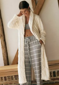 Faux fur morgenkåbe i creme med diamantmønster, der har sjalkrave og lange ærmer. Kombineret med grå stribede loungebukser og en grå top.