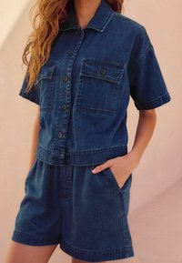 Jeansowa koszula z krótkim rękawem z dwiema kieszeniami na piersi, zapięcie na guziki i dopasowane szorty. Granatowy materiał o gładkiej teksturze.