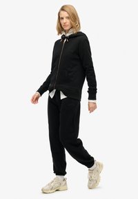 Sudadera negra con cremallera y bolsillos frontales, combinada con pantalones de chándal a juego. El modelo lleva zapatillas beige y una camiseta blanca en capas.