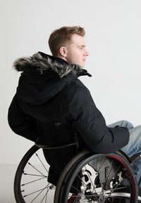 Chaqueta acolchada negra con capucha de piel sintética gris, con cierre de cremallera y puños ajustables, que lleva puesta una persona en una silla de ruedas.