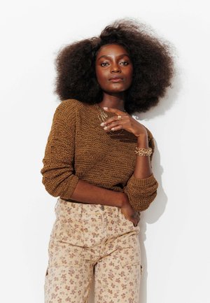 Femme aux cheveux afro naturels portant un pull en maille marron, un pantalon beige à motifs floraux, des bijoux en or, posant avec confiance sur un fond blanc.