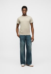 Νεαρός άνδρας στέκεται προς τα εμπρός φορώντας μπεζ κοντομάνικο T-shirt, μπλε τζιν φαρδιάς γραμμής και λευκά παπούτσια μπροστά σε ένα απλό ανοιχτό φόντο.