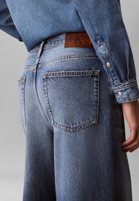 Person trägt hellblaue Jeans und passende Jeansjacke, zeigt Gesäßtasche und braunes Lederetikett mit Calvin-Klein-Logo.