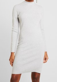 Grå klänning med lång ärm och hög krage. Mjuk stickad tyg, figurnära silhuett, knälång design, minimal detaljer och slät textur.