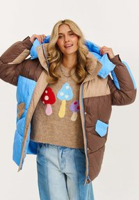 Žena so vlnitými blond vlasmi, oblečená v hnedo-modrom puffriaku cez béžový sveter s farebnými vzormi húb a svetlomodrými džínsami.