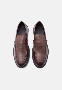 Paire de mocassins marron en cuir à enfiler, avec des détails cousus et des semelles en caoutchouc noir, vus de dessus sur fond blanc.
