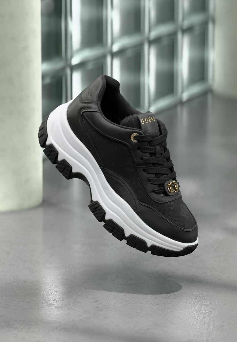 Black athletic sneaker with textured fabric and leather accents. Features a white midsole, rugged black outsole, and gold logo detail.