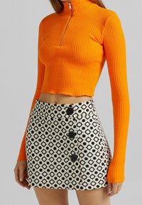 Orange ribbad kort topp med hög hals; parat med en svart och vit mönstrad kjol med cirkulära mönster och svarta knappar.