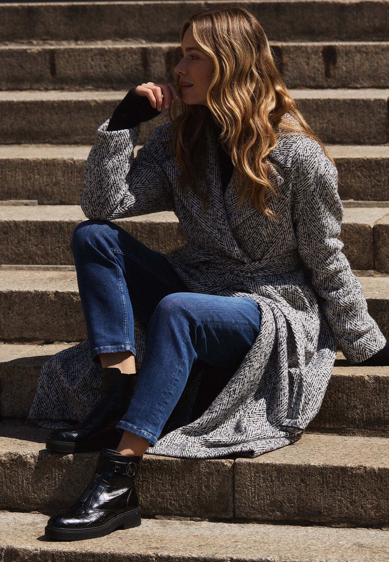 Grijze visgraatjas met een riem, zwarte coltrui, blauwe jeans en zwarte enkellaarsjes, zittend op stenen trappen in natuurlijk licht.