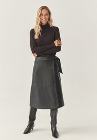 Zwarte leren rok met een gladde textuur, voorzien van een zijstrik en stiksels. Gecombineerd met een donker coltrui en kniehoogte laarzen.