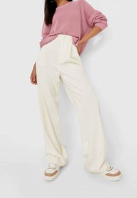 Femme portant un pantalon ample de couleur crème, des baskets blanches et beiges, et un pull en maille rose avec une main dans la poche.