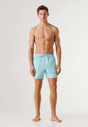 Młody mężczyzna stojący boso, ubrany w jasnoniebieskie kąpielówki w wzory z wiązaniem, na jednolitym tle.