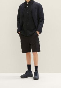 Muž v černé prošívané bundě, černé košili s knoflíky, černých cargo šortkách, černých ponožkách a černých teniskách stojí s rukama v kapsách.