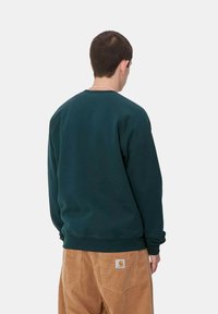 Dunkelgrüner Sweatshirt mit Rundhalsausschnitt und gerippten Bündchen, kombiniert mit beige Cordhosen mit erkennbarer Markenaufnäher auf der Rückseite.