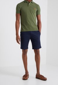 Polo shirt en coton vert avec un petit logo, short marin navy et chaussures bateau marron. La tenue présente une coupe classique et un design simple.