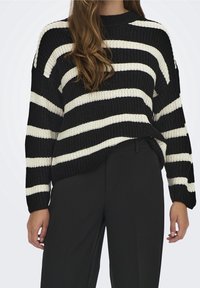 Black and white striped knitted sweater with a relaxed fit, round neck, and dropped shoulders, paired with black, tailored trousers.