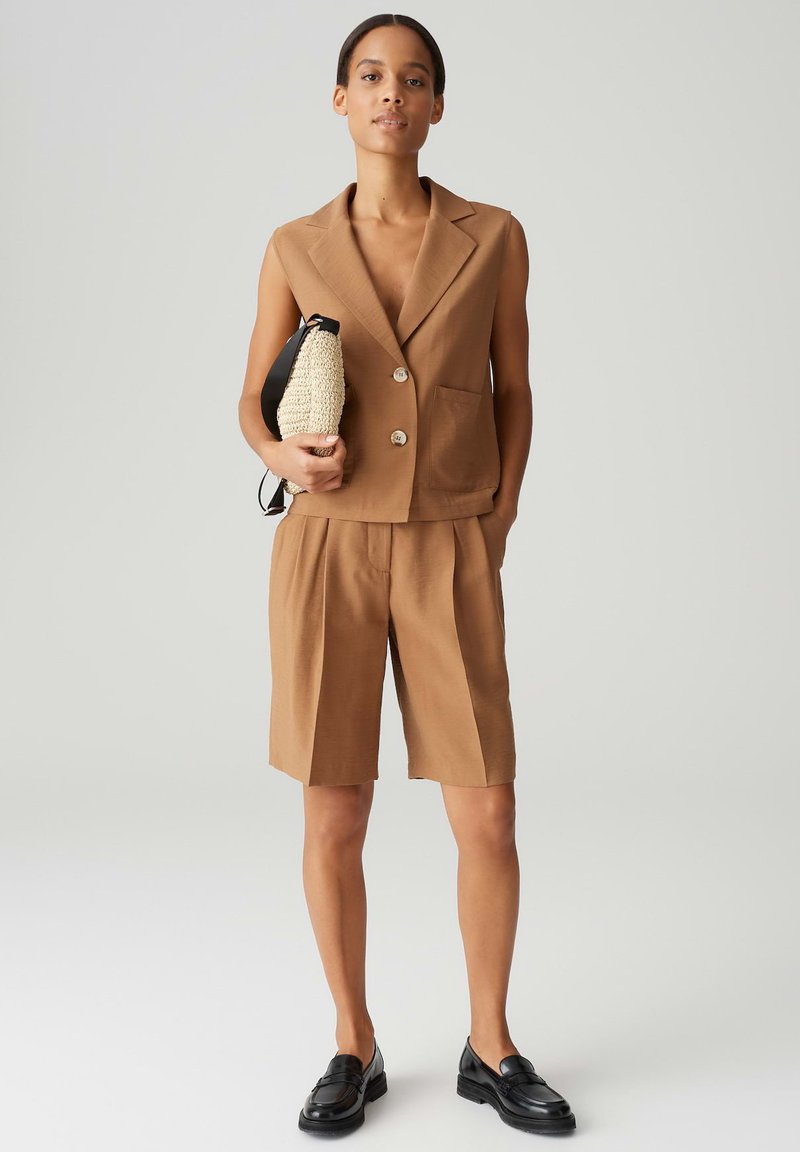 Brauner ärmelloser Blazer und passende knielange Shorts, mit Fronttaschen und lockerer Passform. Strukturiertes schwarzes Clutch und schwarze Slipper.