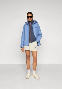 Lichtblauwe waterdichte jas met een oranje rits, gedragen over een donkerblauw shirt en beige korte broek. Gecombineerd met een zonnebril en lichtgekleurde sneakers.