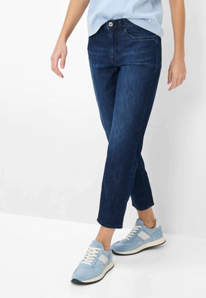 Kvinde iført mørkeblå tætsiddende jeans, lyseblå sneakers med hvide detaljer og en lyseblå skjorte, stående foran en hvid baggrund.