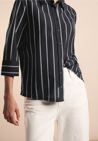 Chemise noire à boutons avec des rayures verticales blanches, affichant une coupe décontractée et des manches retroussées, associée à un pantalon blanc texturé.