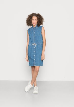Vestido camiseiro sem mangas em denim azul, com frente abotoada, cintura com cordão e corte recto. Combinado com ténis brancos.