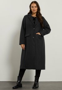 Kobieta w długim czarnym płaszczu z guzikami i kieszeniami, czarnych spodniach oraz czarnych botkach za kostkę, stojąca na tle jednolitego jasnego tła.