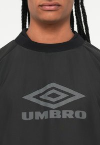 Sõrmustega juukseid kandva isiku seljas on must Umbro särk, millel on rinnal suur hall kahekordne rombikujutis ja brändinimi.