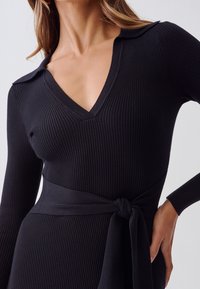 Svart ribbad klänning med V-ringning och krage, med långa ärmar, knytbar midja och en mjuk textur. Justerbar passform genom bältet.