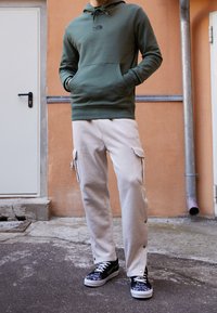 Felpa verde oliva con tasca frontale, abbinata a pantaloni cargo beige con tasche laterali e chiusure a scatto, completata da sneaker nere.