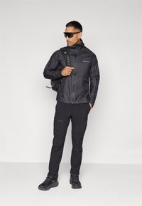 Črna lahka jakna z zadrgo, kapuco in logotipom, v kombinaciji s usklajenimi hlačami. Dodatki vključujejo nahrbtnik in sončna očala.