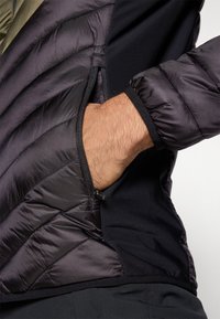 Roka ieliktas melnā quilted un softshell jakas sānu kabatā ar rāvējslēdzēju, redzams elastīgs aproces un auduma tekstūra.