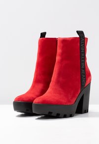 Botas de tobillo de ante rojo con tacones y suelas gruesas negras, con tiradores negros que tienen el texto "Calvin Klein Jeans" en el lateral.
