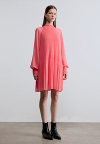 Vestito fluente fino al ginocchio in rosa acceso, caratterizzato da collo alto, maniche lunghe, texture plissettata, abbinato a stivaletti neri.