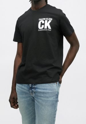 Homme portant un t-shirt noir Calvin Klein CK et un jean bleu clair, debout avec une main dans la poche devant un fond uni.