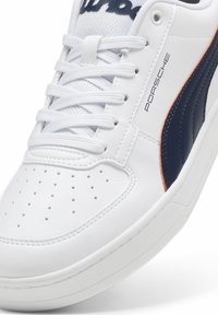 Baskets en cuir blanc avec lacets blancs, logo Puma noir, texte "Porsche" sur le côté et zone perforée au niveau des orteils.