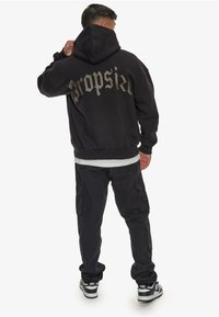 Schwarzer Kapuzenpullover mit großem, stilisiertem "propsize"-Text auf dem Rücken. Kombiniert mit schwarzen Cargo-Hosen und Sneakers mit weißer Sohle.
