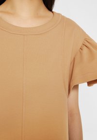 Haut beige à manches courtes en matériau doux, avec un col rond et des accents froncés sur les manches avec des détails de couture visibles.